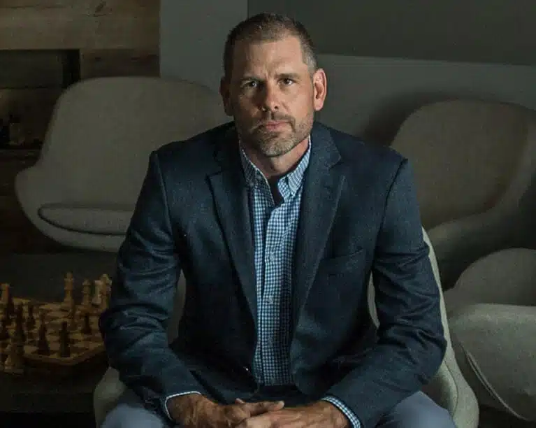 Scott Hermann, the CEO of IDIQ, wearing a dark blazer and a checkered shirt, sits in a modern room. Behind him, there is a chessboard on a table and two cushioned chairs. The lighting is dim, highlighting his serious expression related to credit management.