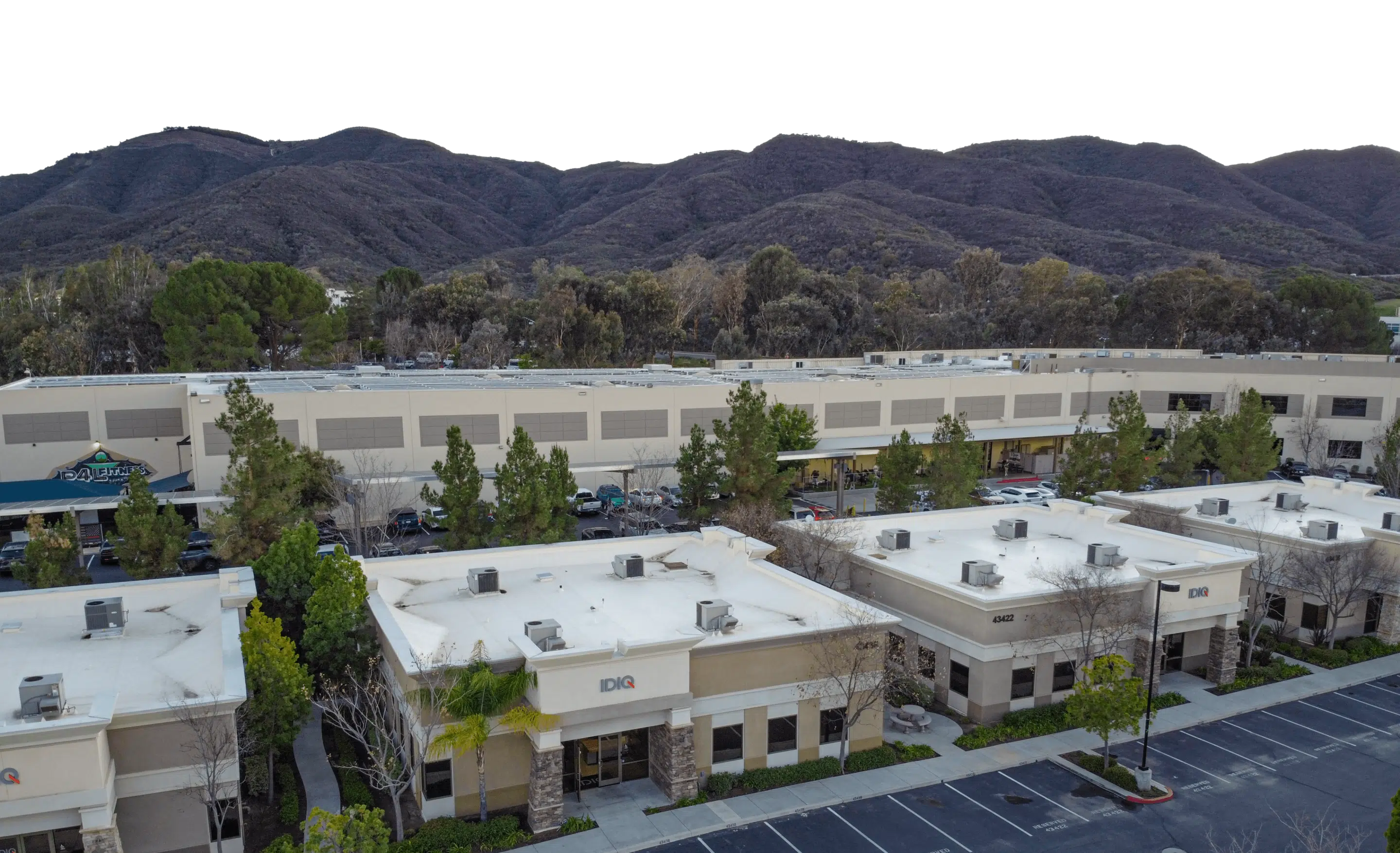 An aerial view of a commercial complex with several buildings and parking spaces in the foreground, against a backdrop of rolling green hills and mountains.