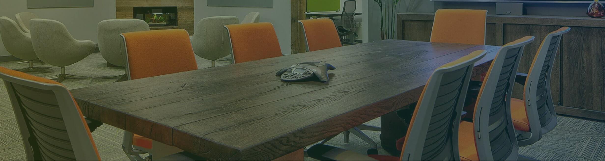 A modern conference room features a rustic wooden table surrounded by eight chairs with orange and black upholstery. In the background, there is a cozy seating area with plush gray chairs and a fireplace. A conference phone is placed in the center of the table, emphasizing discussions on credit management.