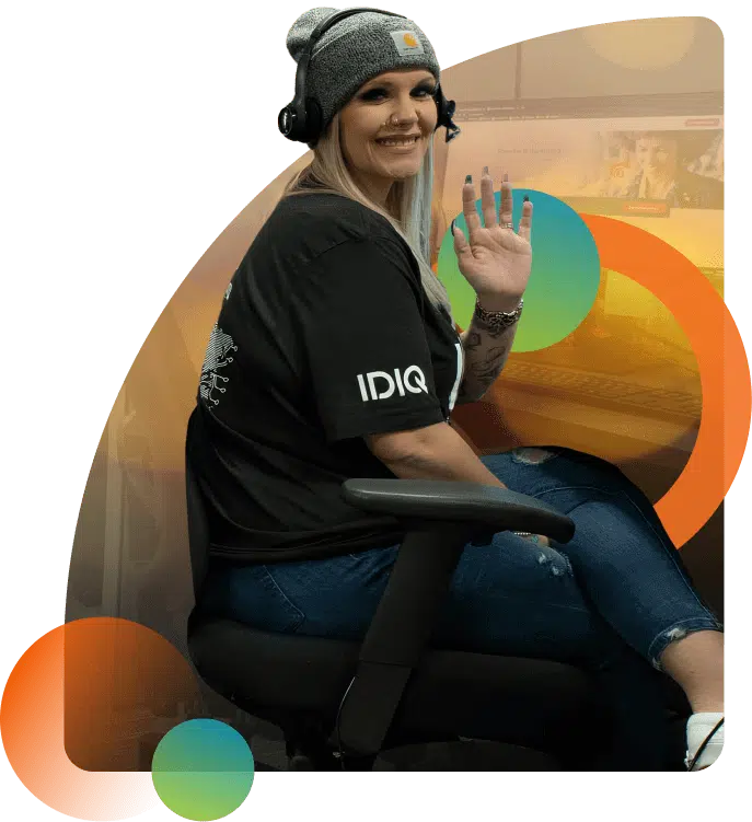 A woman with long blonde hair wearing a black shirt and a gray beanie, sitting in a swivel chair with a headset. She is smiling and waving, with colorful circular graphics in the background.