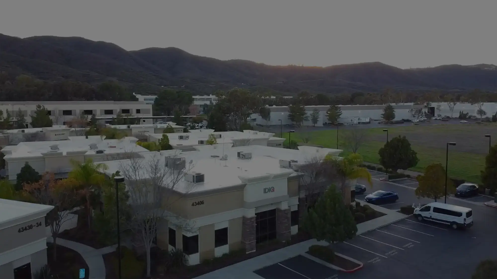 An aerial view of a commercial area with several single-story buildings and a parking lot. In the background, green fields and hills are visible under a twilight sky.