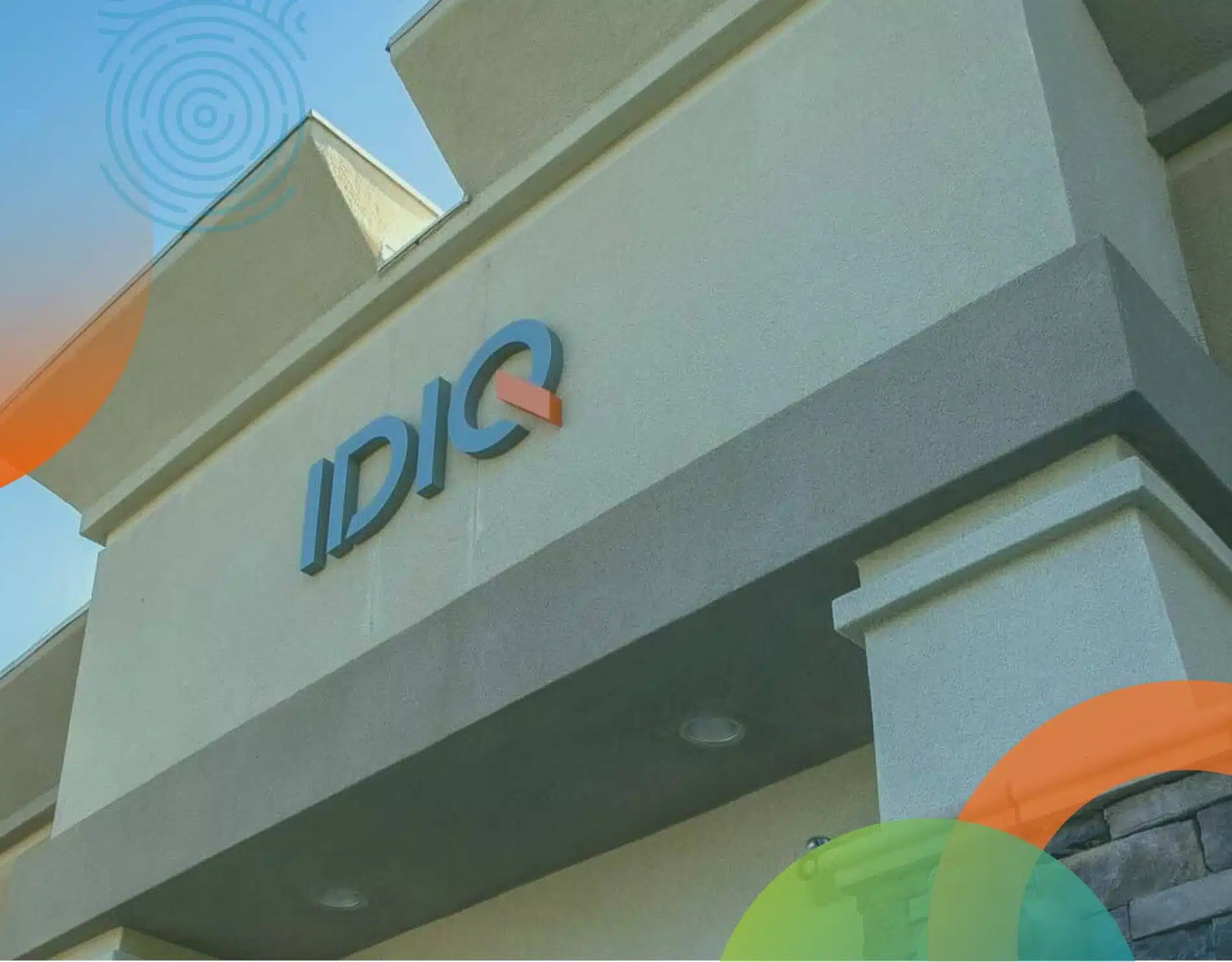 The exterior of a building featuring the sign "IDIQ" in blue and orange colors. The structure is light-colored with a modern architectural design. Colorful circular overlays appear in the bottom corners.