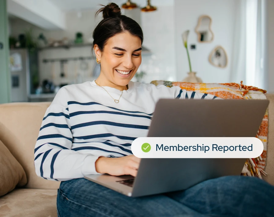 Young woman sitting on her couch at home on her laptop smiling with a notification popping out that says Membership Reported