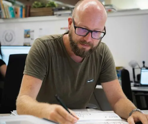 A man with glasses and a beard is seated at a desk, focused on his work.
