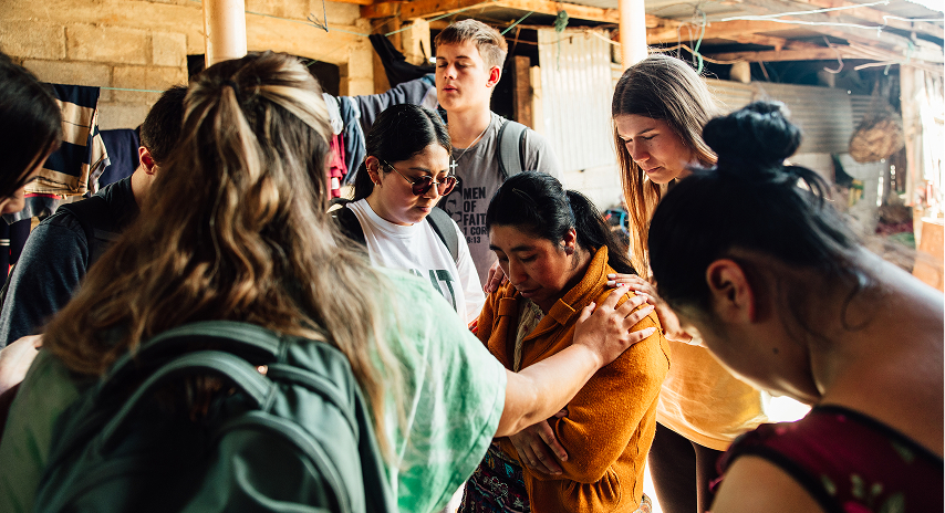 A small group of people gathers around a woman, offering support and prayer during an outreach or mission activity in a rural setting.