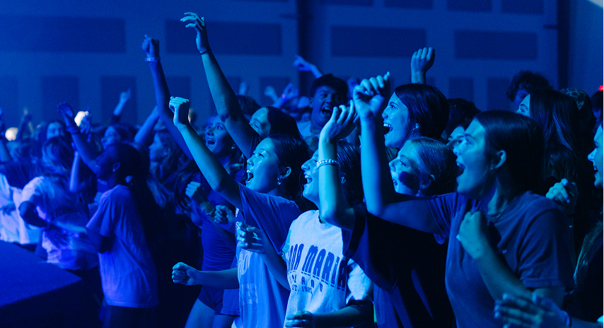A crowd of energetic teenagers cheers and raises their hands during a lively youth worship event with blue stage lighting.