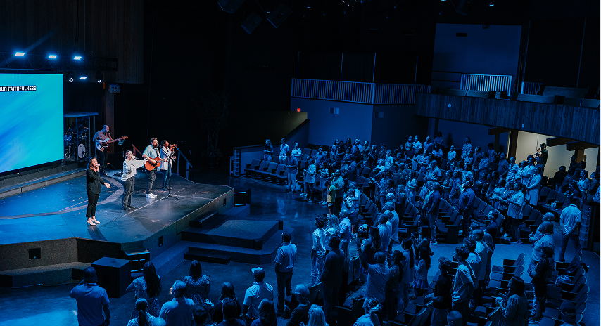A worship band leads music on stage while a large congregation stands and participates during a church service in a modern auditorium.