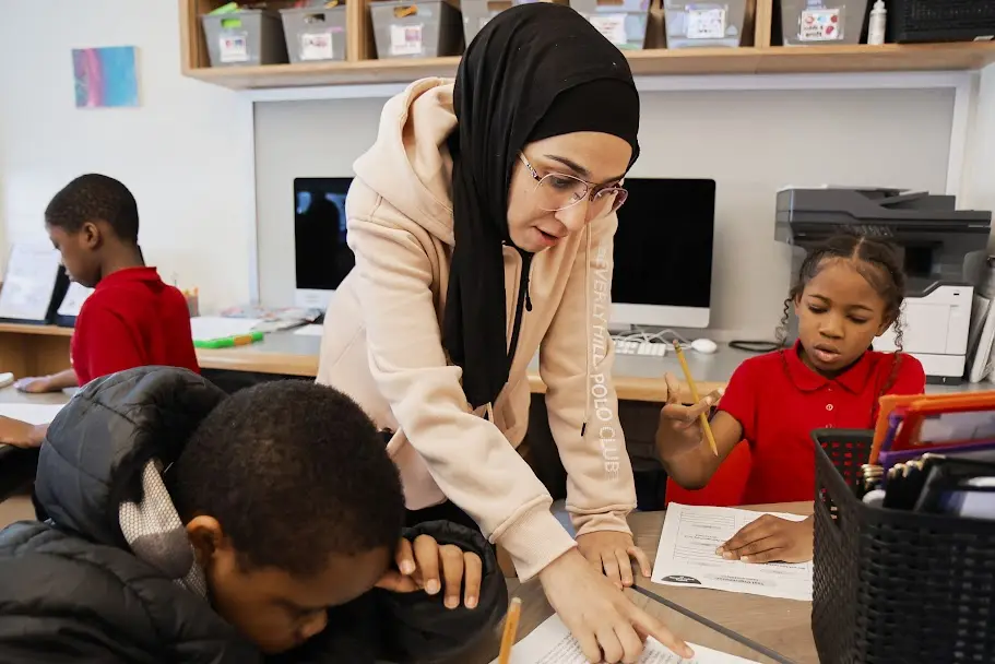 Teacher leaning over to help two elementary students with classwork