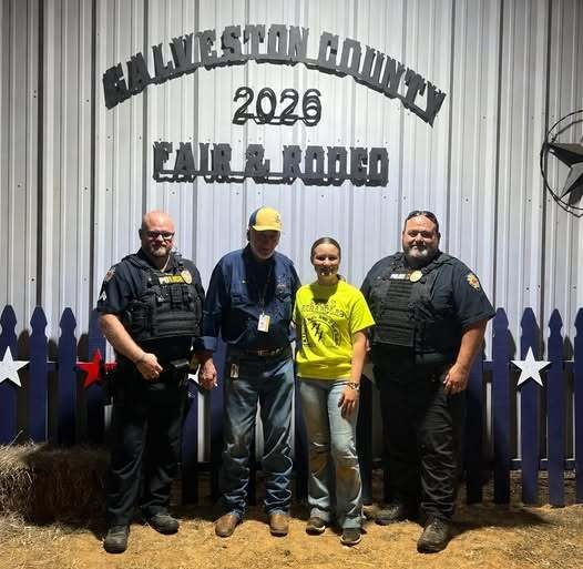 Four people standing in front of a metal wall with a sign reading 'Hallam Livingston County 2026 Fair & Rodeo', two men wearing police uniforms and one man in a blue shirt and cap, and a woman in a yellow t-shirt and jeans.