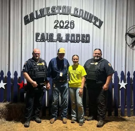 Four people standing in front of a metal wall with a sign reading 'Hallam Livingston County 2026 Fair & Rodeo', two men wearing police uniforms and one man in a blue shirt and cap, and a woman in a yellow t-shirt and jeans.