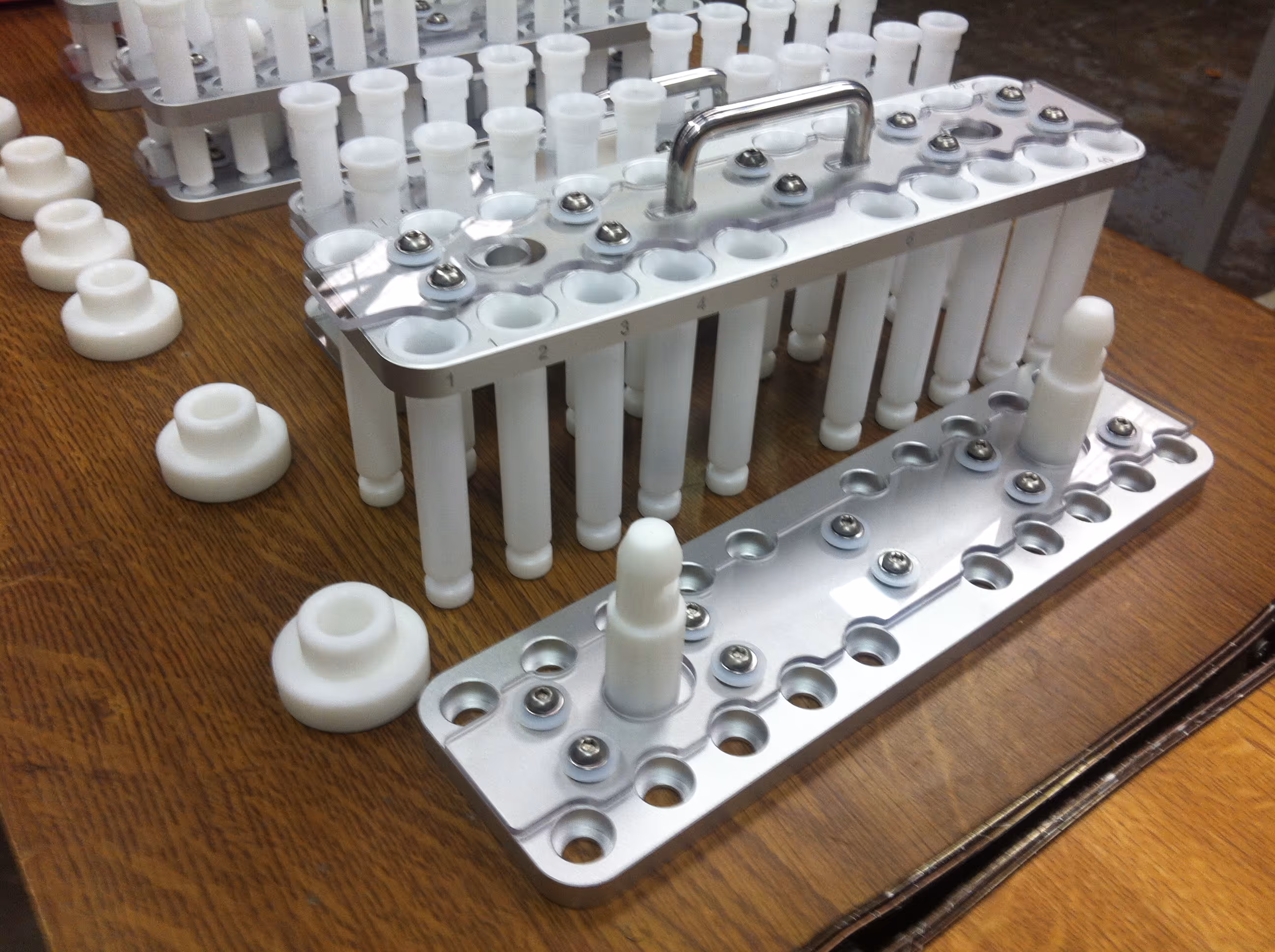 Custom aluminum and plastic fixture with numbered slots and cylindrical components, likely used for precision assembly or testing, placed on a wooden workbench.