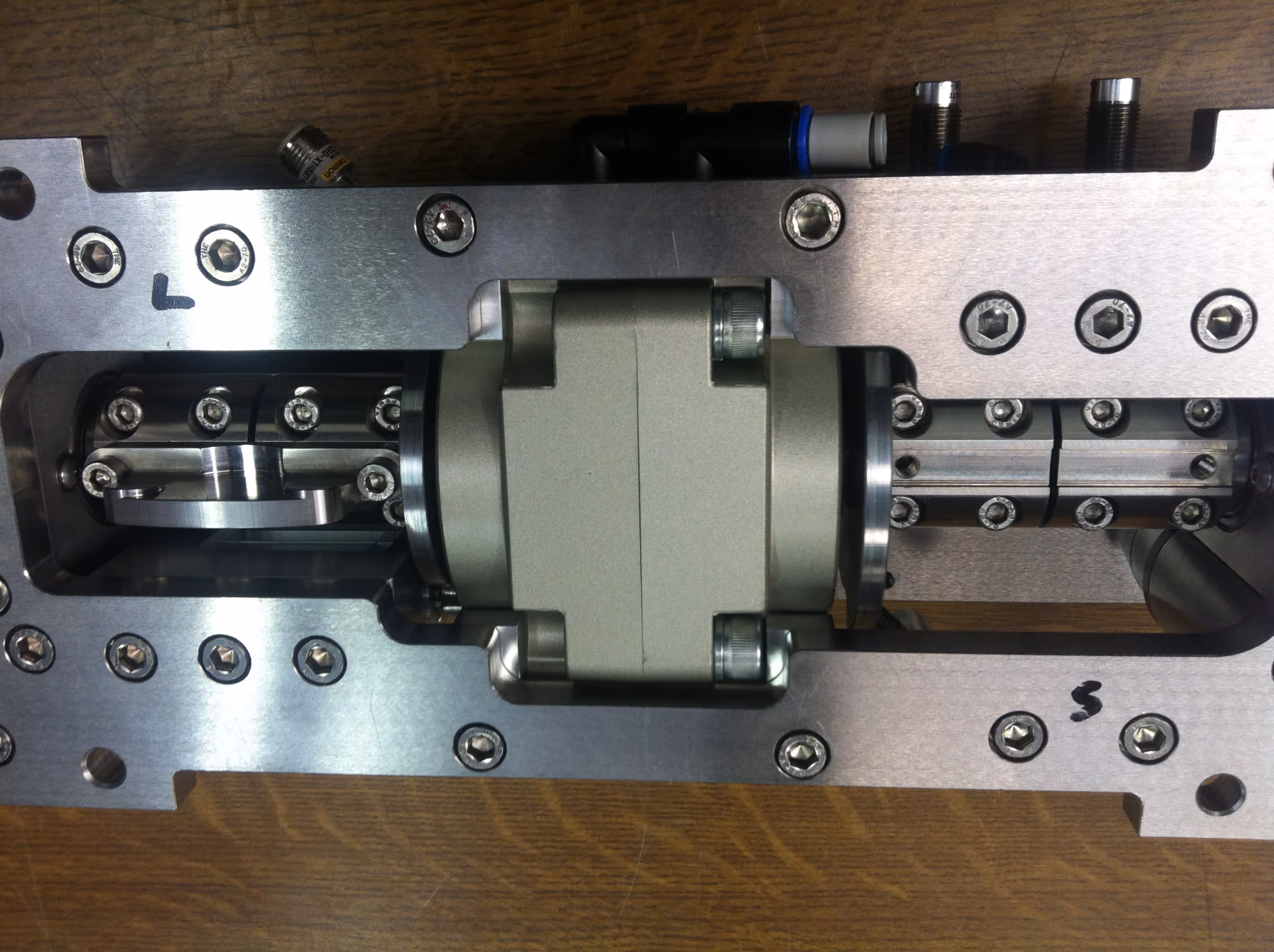Precision-engineered metal assembly with a pneumatic or hydraulic actuator mounted within a machined aluminum frame, secured by multiple bolts on a wooden surface.