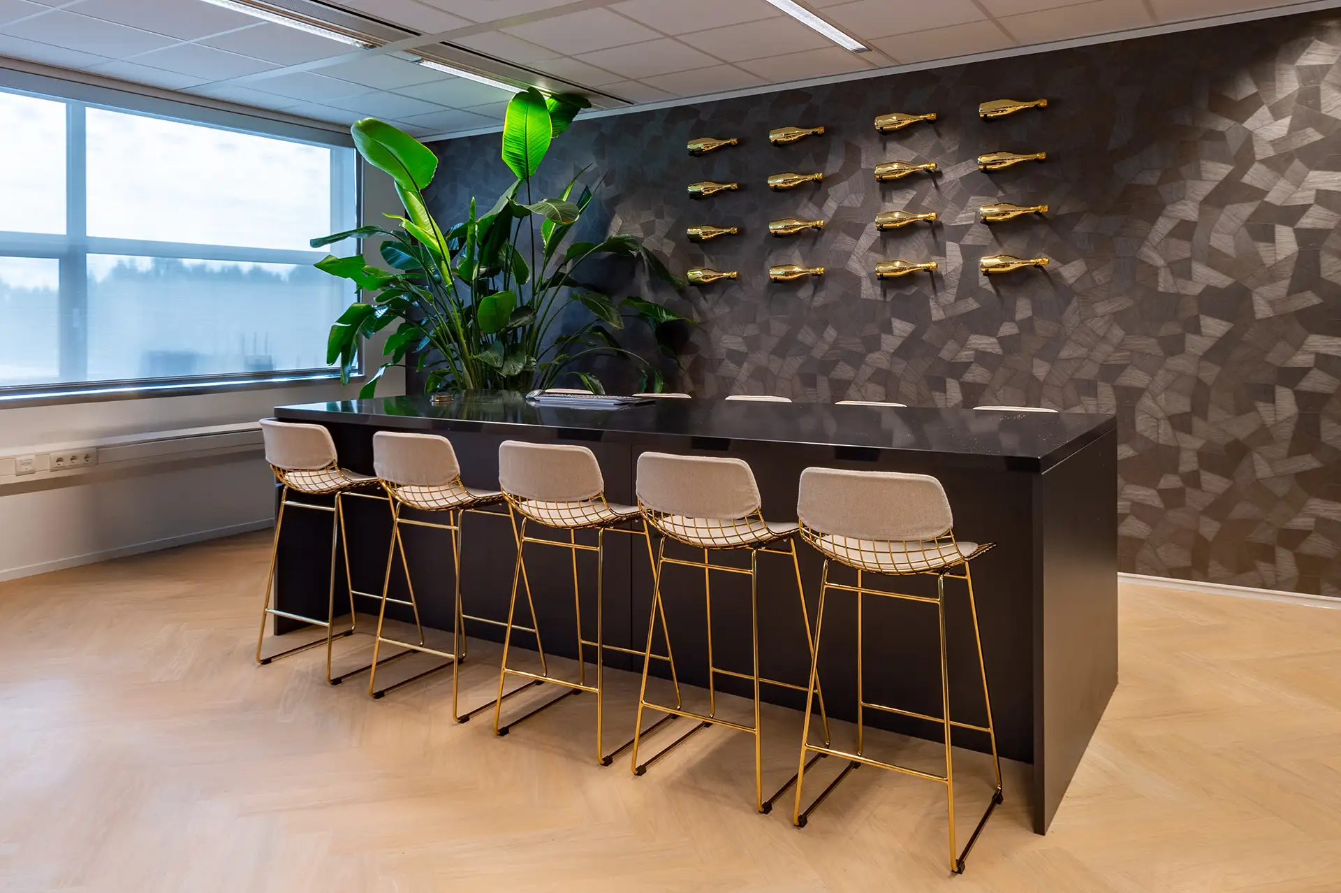 Moderne kantoorruimte met een zwarte bar, zes hoge stoelen met gouden frames, grote groene planten en een muur met gouden decoratie.
