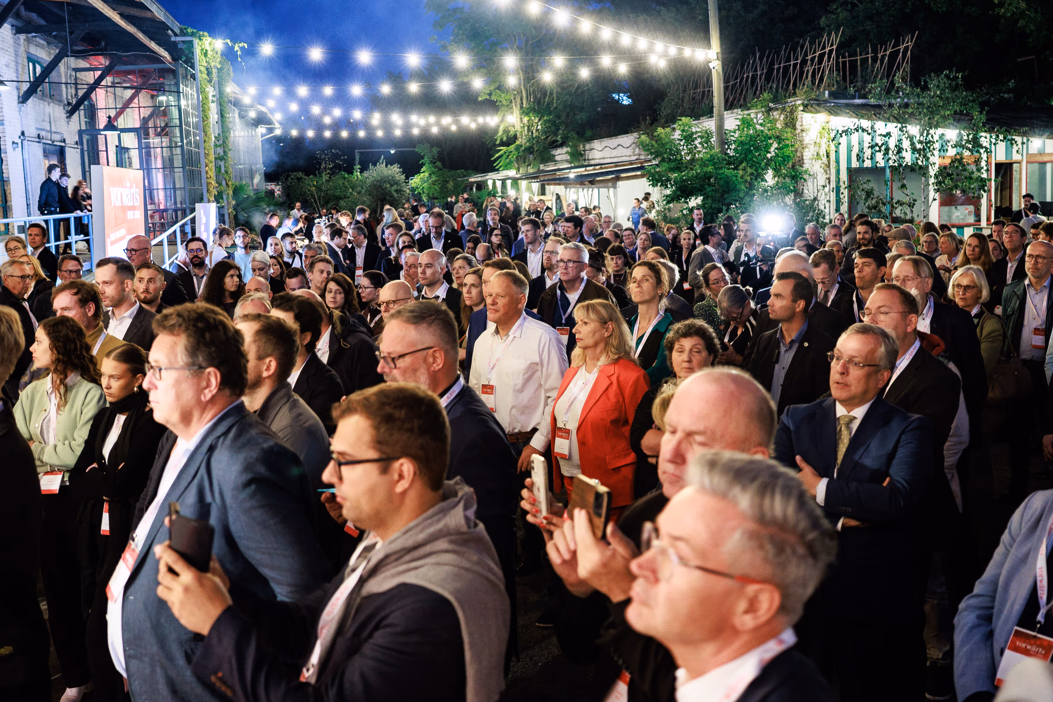 Publikum steht unter Lichterketten im Freien und verfolgt aufmerksam einen Fachvortrag. Veranstaltungskonzeption und Eventplanung – ASK 