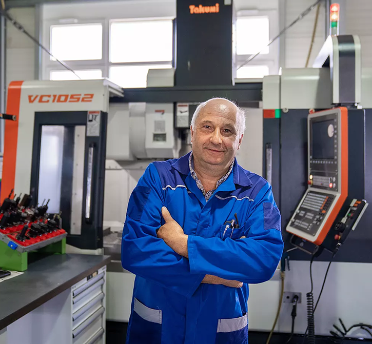 Man wearing a blue work uniform standing with arms crossed in a workshop in front of a Takumi CNC machine.