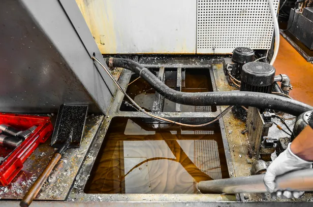 Industrial machine platform with pools of brownish liquid, hoses, and a gloved hand holding a metal rod.