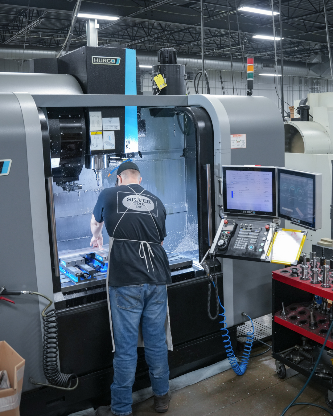 Worker operating a Hurco CNC milling machine in a factory workshop with tool racks nearby.