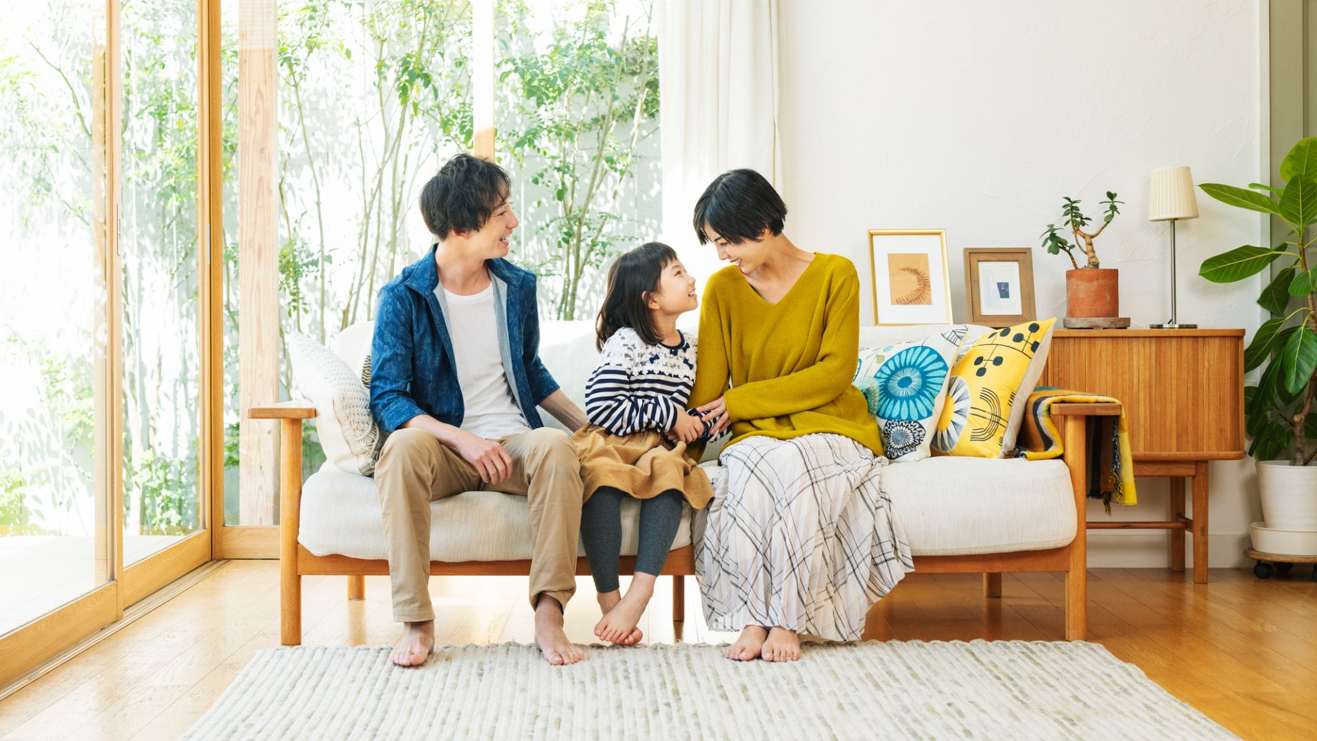 A family of three (man, woman, and young girl) are sitting barefoot on a couch in a bright, plant-filled room, seemingly interacting.