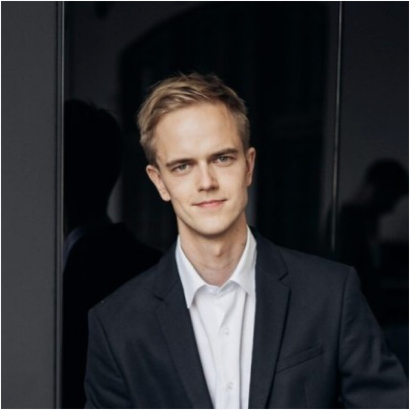 Young man with blond hair wearing a dark suit jacket and white shirt, smiling slightly against a dark background.