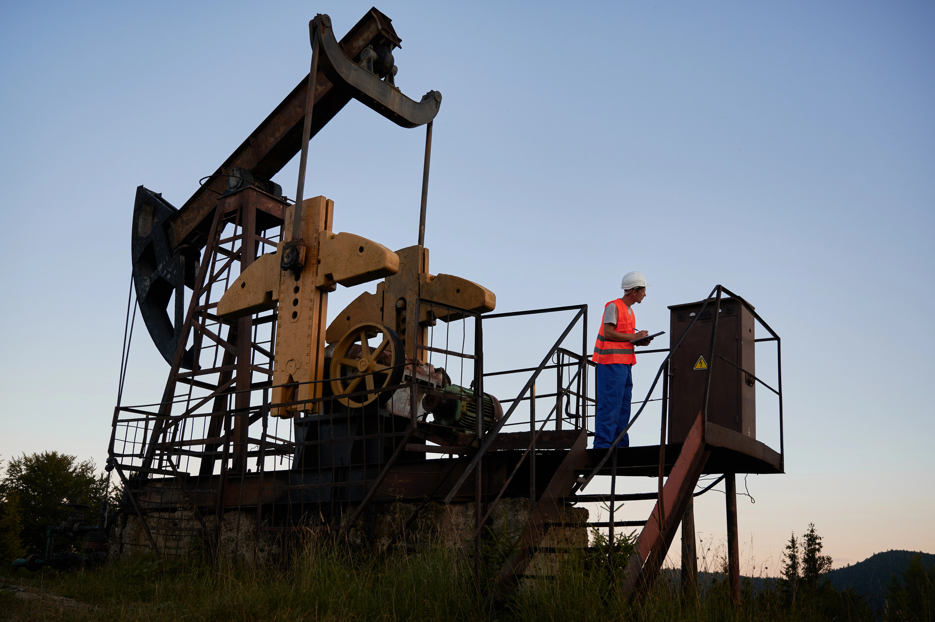 Worker in safety gear inspecting a large oil pump jack at an outdoor site during dusk.