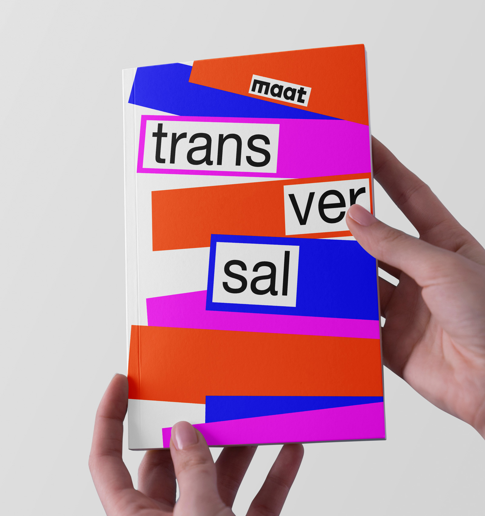 Person holding a white book cover decorated with bold orange, blue, and pink geometric shapes and words 'maat trans versal' in black.