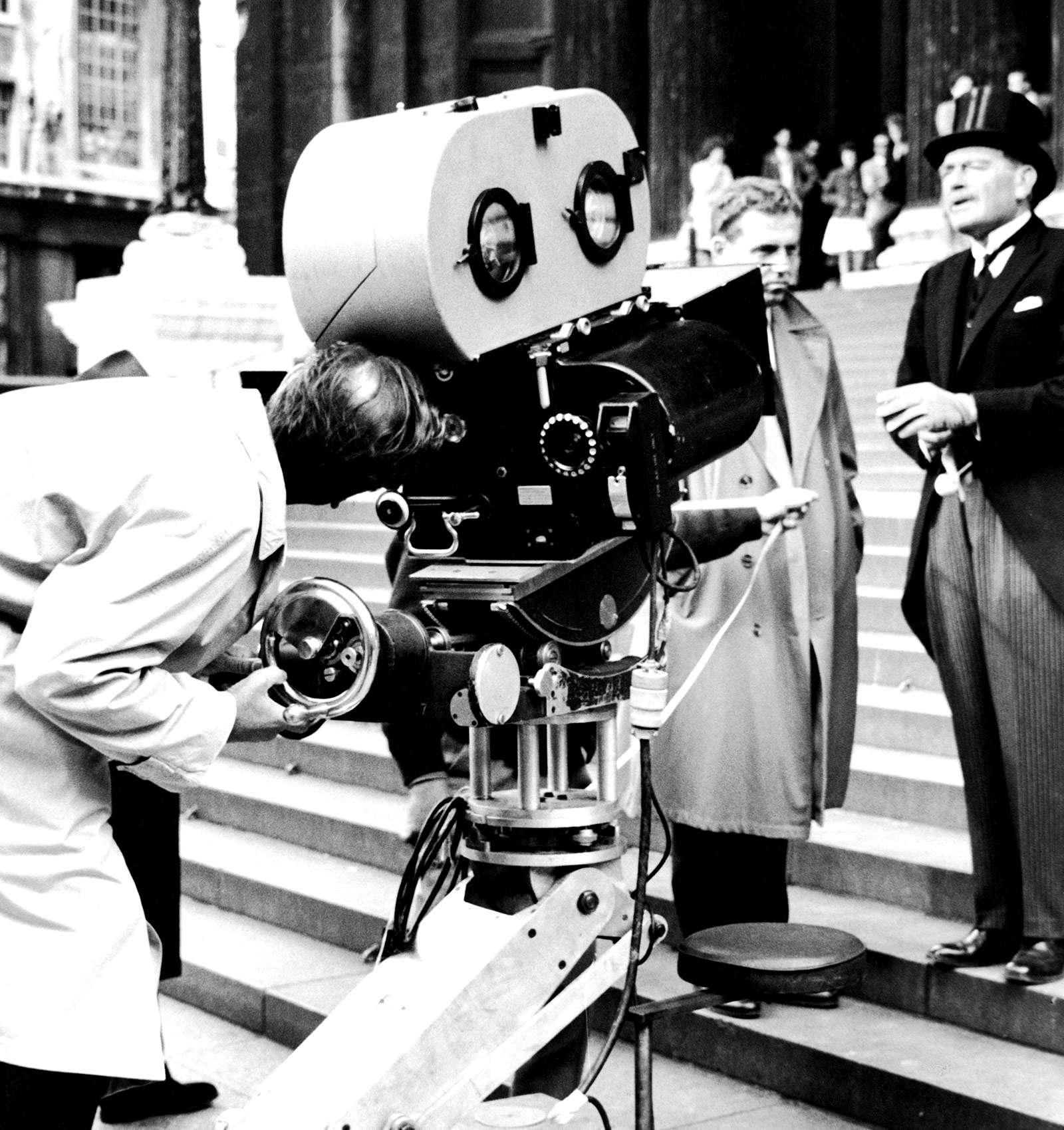 Black and white photo of a film crew operating a large vintage movie camera in front of stone steps, with two men in period clothing standing nearby.