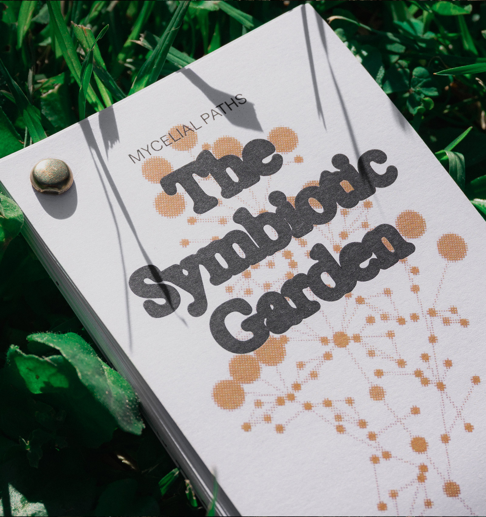 Close-up of a card titled 'The Symbiotic Garden' with the subtitle 'Mycelial Paths' featuring an abstract pattern of connected orange dots and lines, lying on green grass.