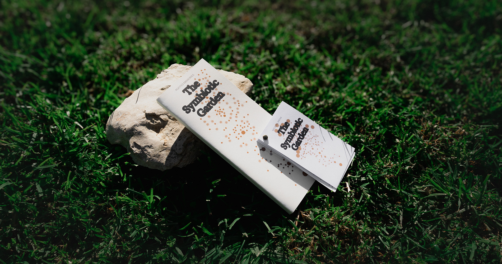 Two booklets titled 'The Symbiotic Garden' placed on a light-colored stone on green grass.