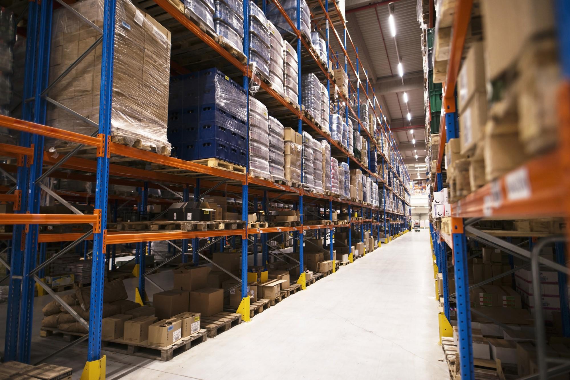 Long aisle in a warehouse with tall blue and orange shelving filled with wrapped pallets and cardboard boxes.