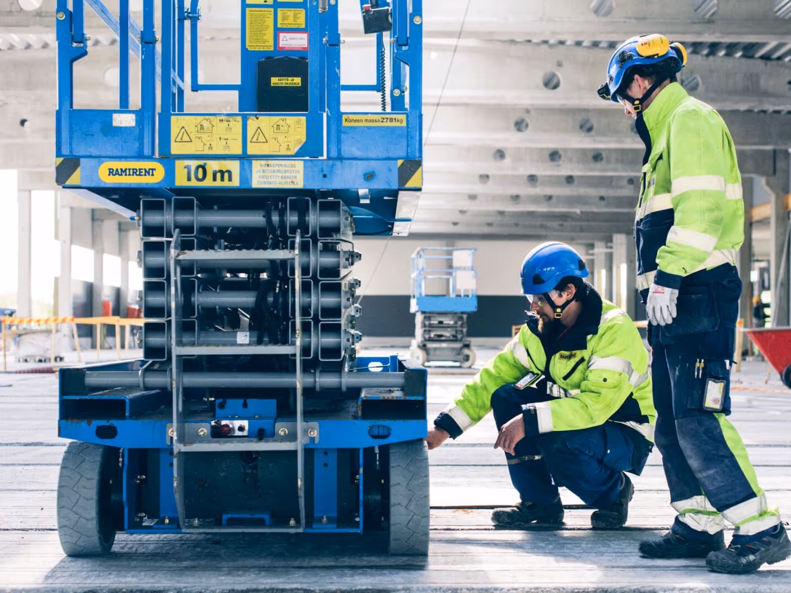 Two construction workers in high-visibility yellow jackets and safety helmets inspecting a blue Ramirent aerial lift platform. One worker is kneeling to examine the base of the equipment while the other stands watching.
