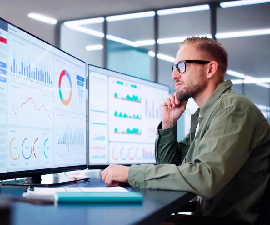 A professional man wearing glasses and a green shirt sits at a modern office desk, analyzing data on multiple large computer screens displaying charts, graphs, and analytics dashboards.