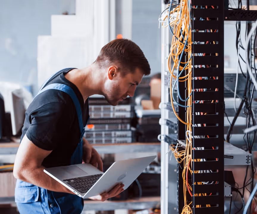 A technician wearing blue overalls and a black t-shirt is inspecting a server rack filled with tangled network cables. He is holding a laptop and leaning forward, closely examining the connections in a data center or server room.