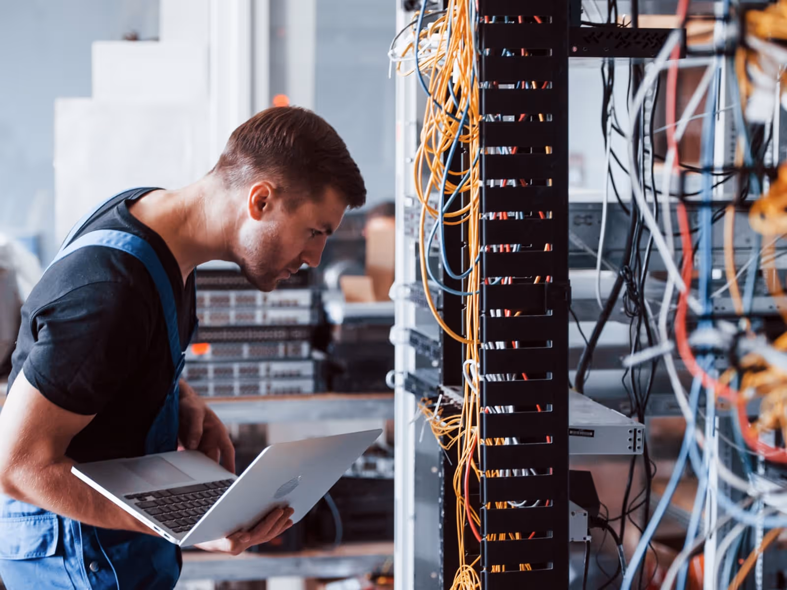 Un técnico vestido con un mono azul y una camiseta negra inspecciona un rack de servidores lleno de cables de red enredados. Sostiene una computadora portátil y se inclina hacia adelante, examinando detenidamente las conexiones en un centro de datos o una sala de servidores.