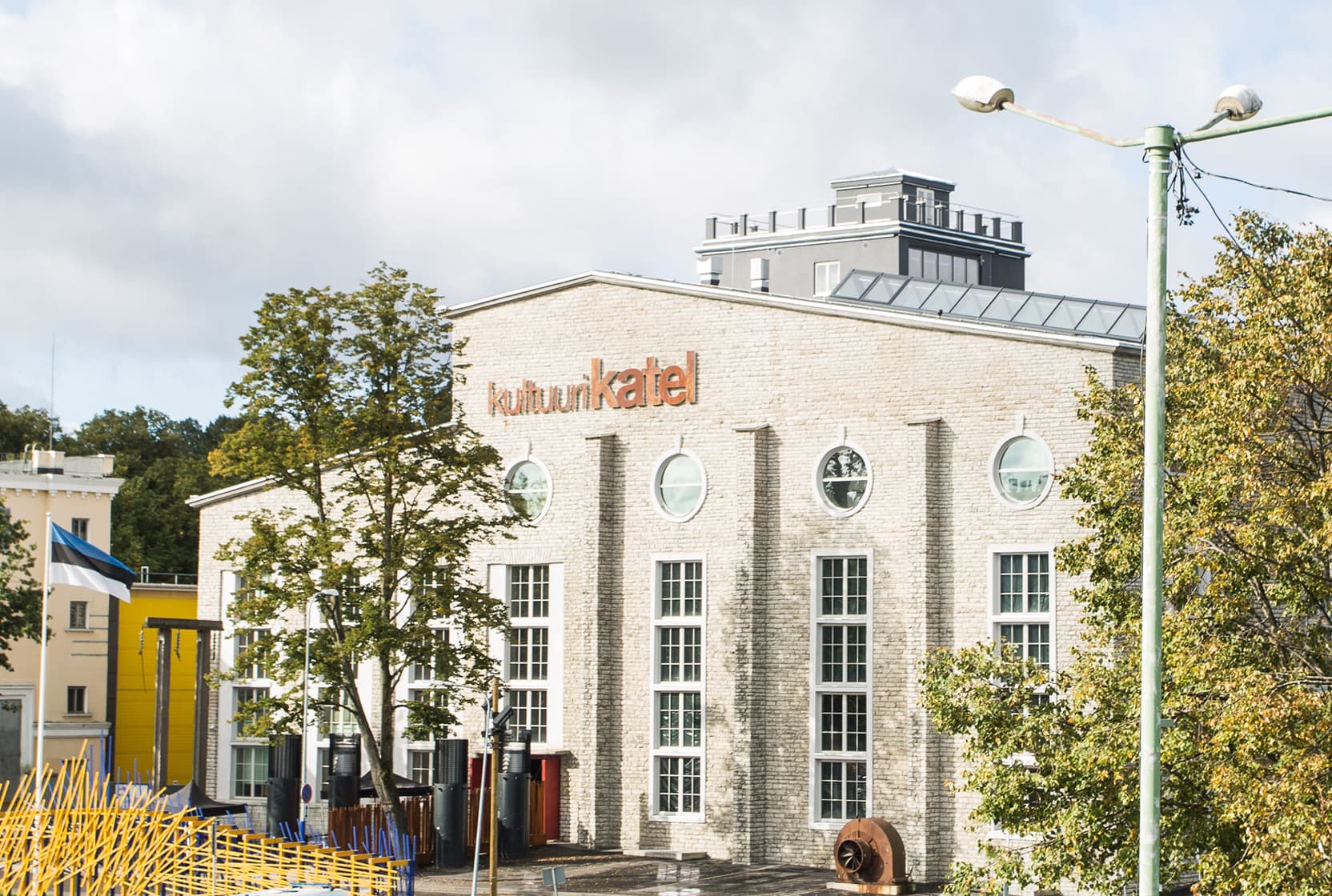 Modern light-brick building labeled “kultuurikatel,” with large round windows, a tree on the left, streetlight on the right, and partly cloudy sky; urban scene with a yellow building in the background.