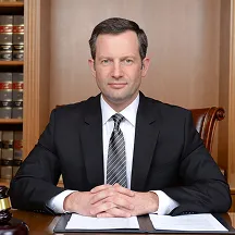 Man in a black suit and striped tie sitting at a desk with folded hands, with bookshelves in the background.