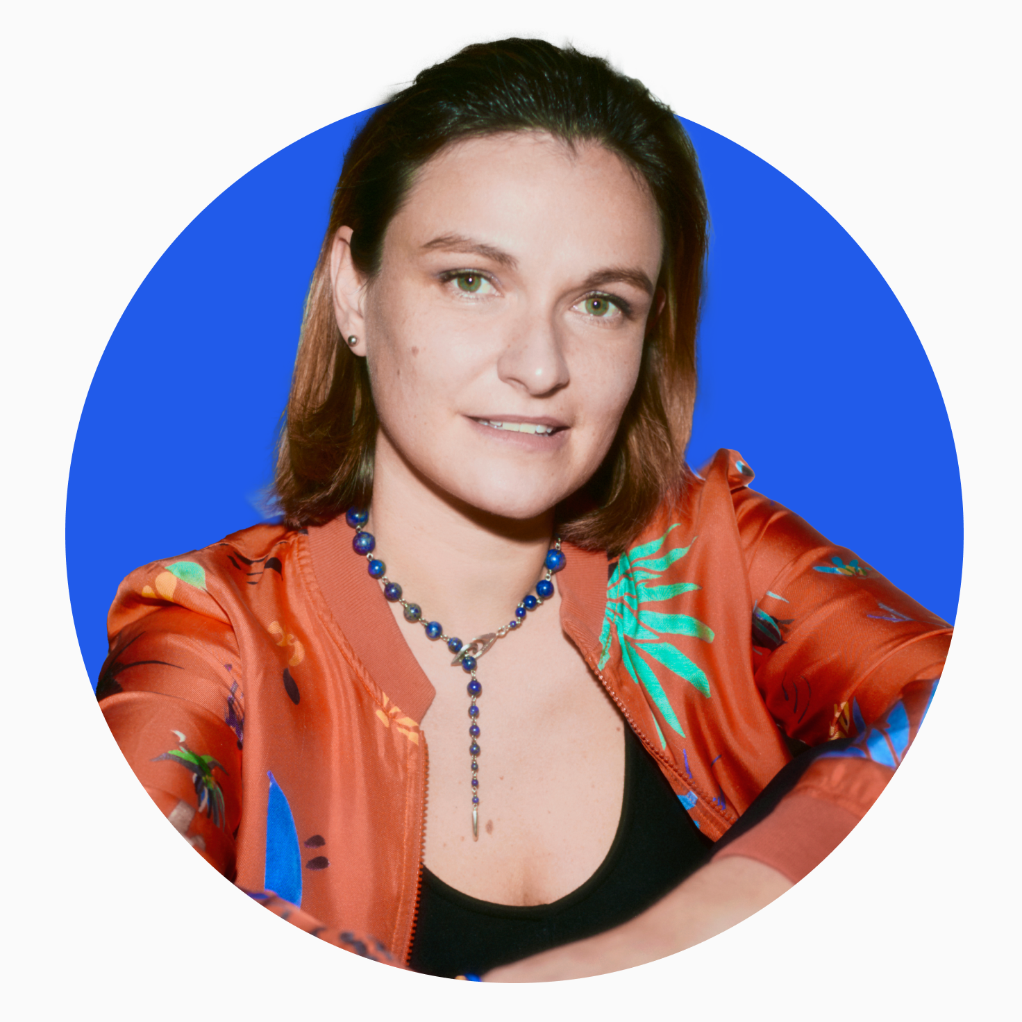 Smiling woman with light skin, green eyes, wearing a colorful orange jacket and blue beaded necklace against a blue circular background.