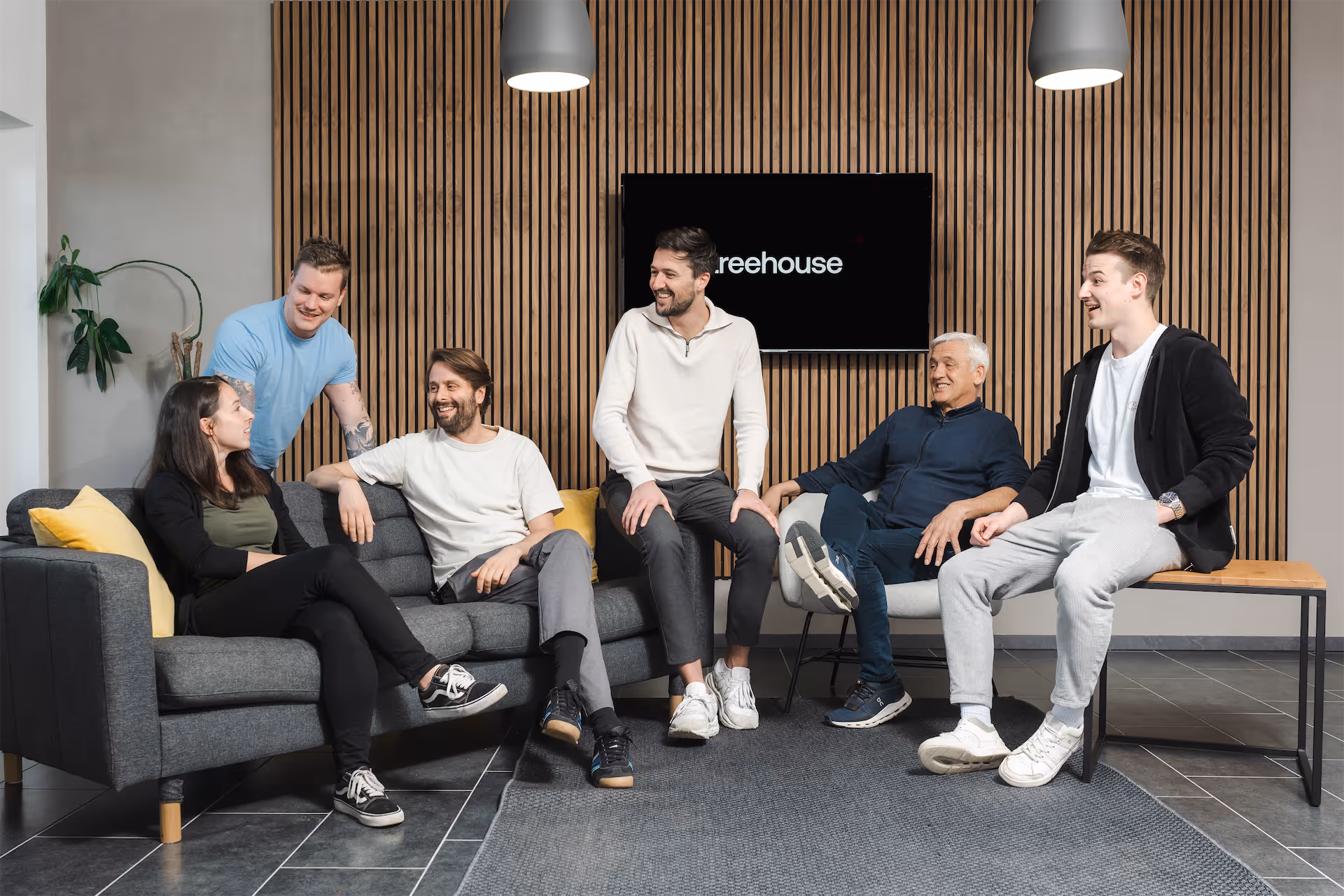 A group of six people gathered in a modern office lounge area around a gray sectional sofa. The backdrop features a wooden slat wall with a mounted TV displaying 'reehouse' branding. The casual, friendly team setting includes both seated and standing individuals in a bright, contemporary workspace with natural lighting