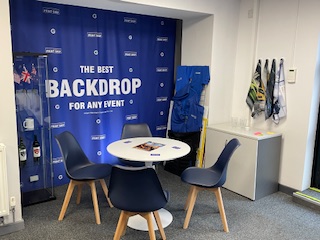 Corner of our showroom with a small round white table surrounded by three blue chairs for customers, and a blue backdrop displaying 'THE BEST BACKDROP FOR ANY EVENT' text.