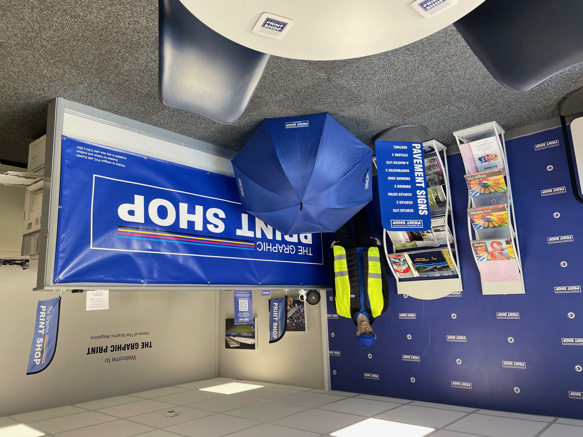 Interior of The Graphic Print Shop with a reception desk, sample banners, a mannequin dressed to demonstrate our corporate clothing range, and a blue personalised umbrella on the floor.