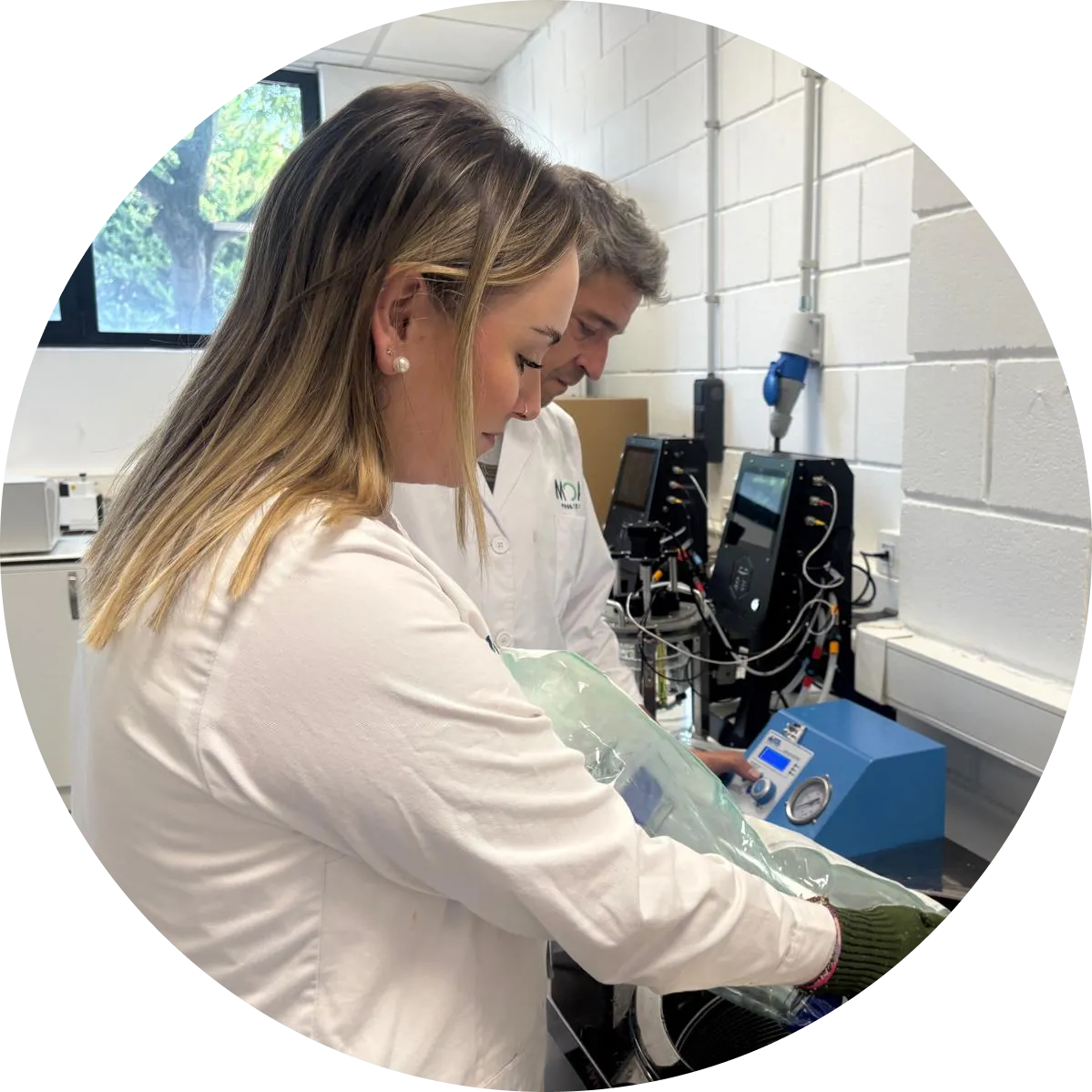 Female MOA researcher in white lab coat examining green biological sample or algae culture while male colleague works in background in modern laboratory with scientific equipment, presented in circular frame design