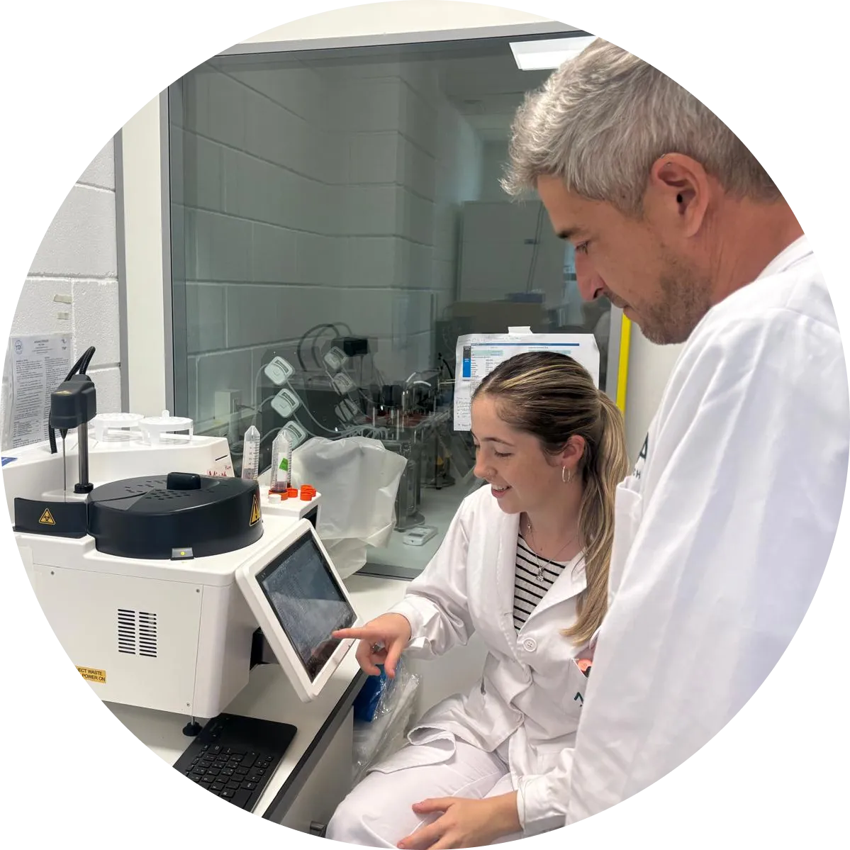 Senior male MOA scientist with gray hair supervising female researcher operating advanced laboratory analysis equipment with digital touchscreen interface in modern research facility, presented in circular frame design