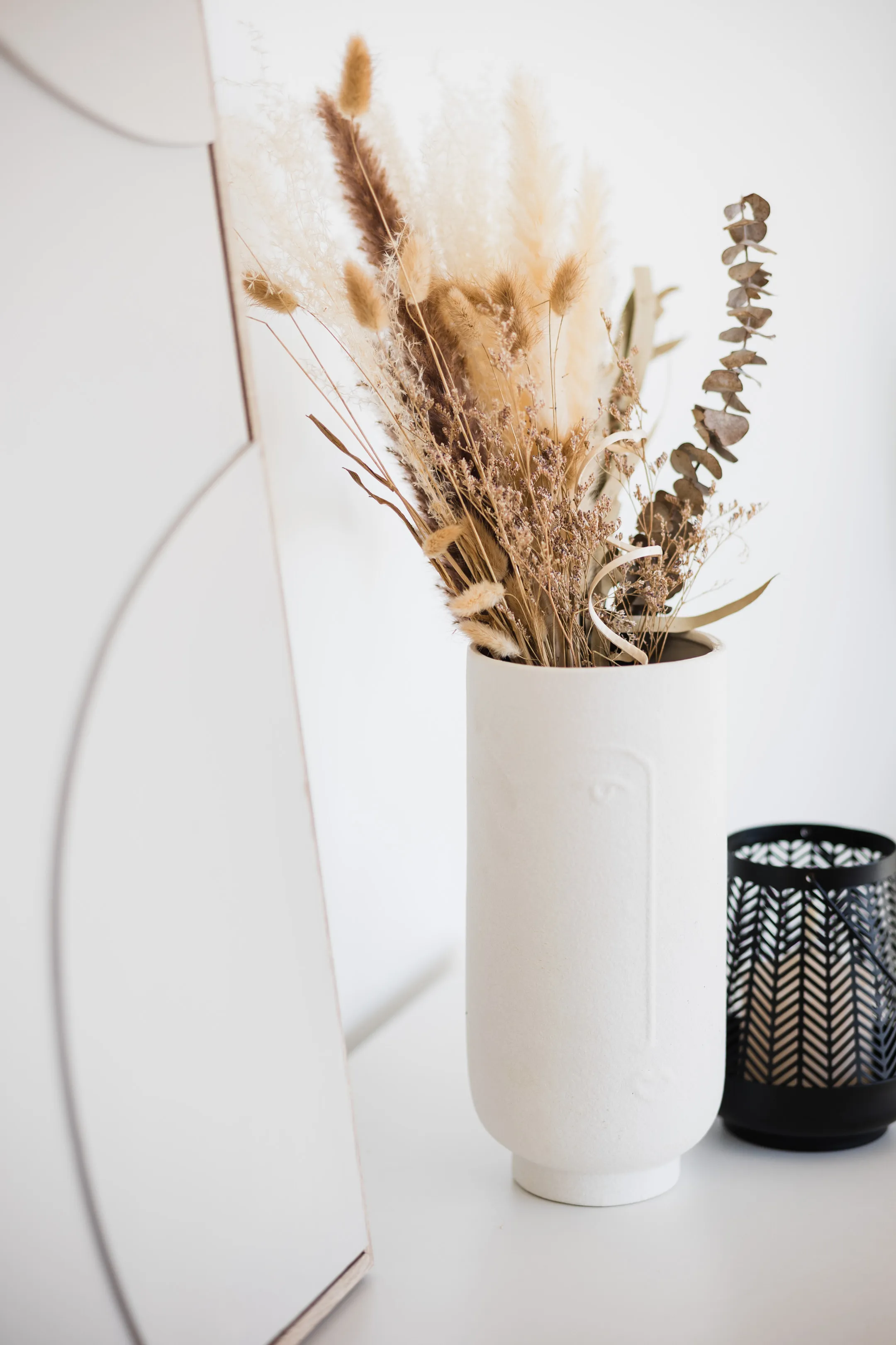 Exzellente Produktfotografie von Visual Artisan für Heimtextilien und Dekoration. Stilvolles Arrangement aus einer weißen Designvase mit getrockneten Gräsern und einem schwarzen Windlicht, professionell inszeniert für Onlineshops und Marketingmaterialien. Ihr Partner für beeindruckende Produktvisualisierungen.