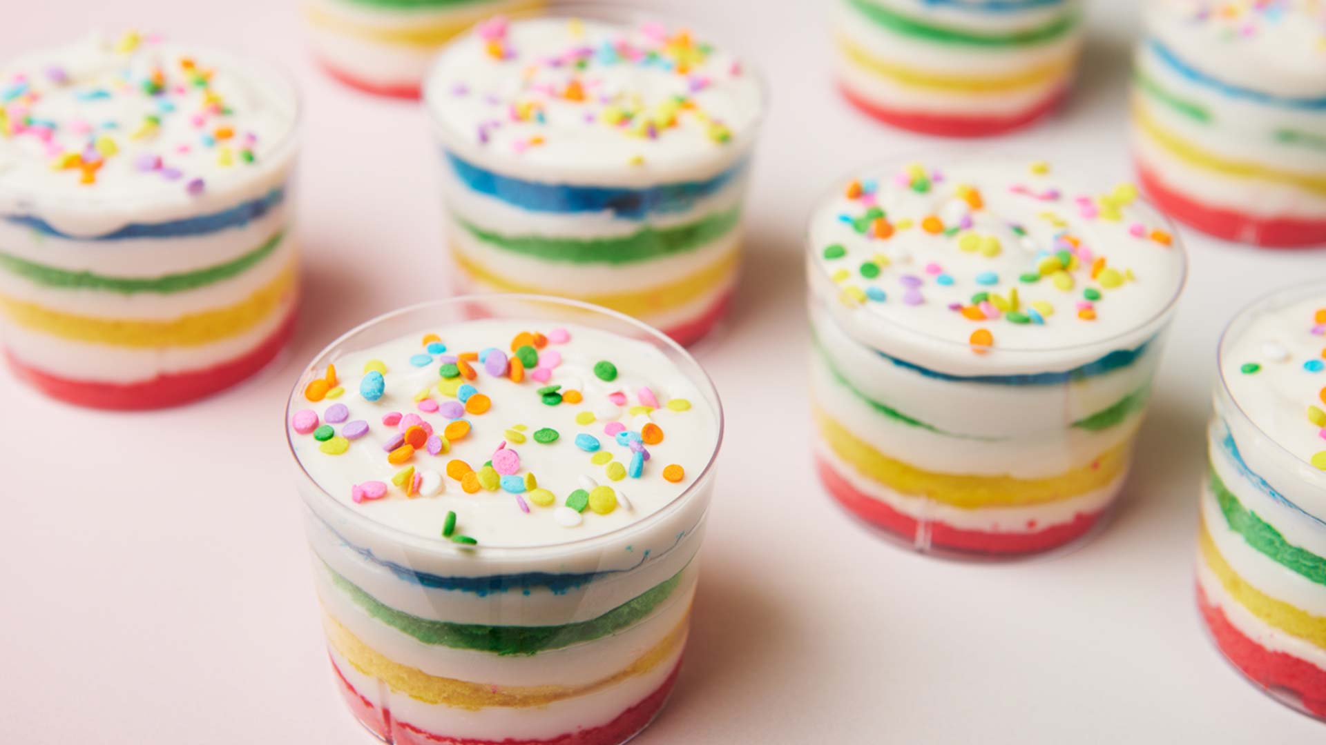 Mini rainbow cake stacks layered with red, yellow, green, blue, and white frosting, topped with colorful sprinkles