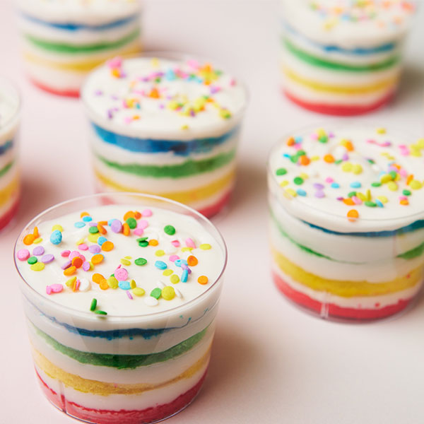 Mini rainbow cake stacks layered with red, yellow, green, blue, and white frosting, topped with colorful sprinkles and arranged on a clean white surface for a Pride-themed dessert display.