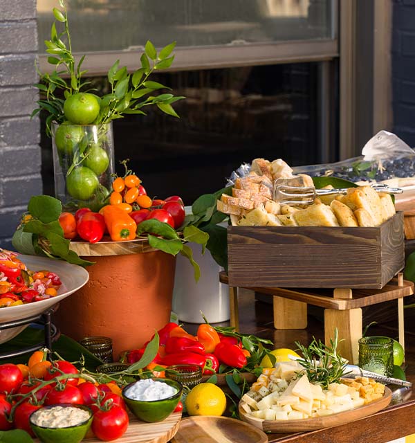 Summer catering spread with fresh tomatoes, peppers, berries, bread, cheese, citrus, herbs, and dips arranged on a wooden buffet table.
