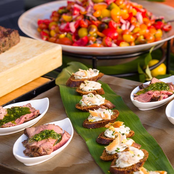 Assorted catered rooftop food display with sliced steak and herb sauce in white tasting bowls, crostini with creamy topping on a banana leaf, fresh tomatoes, peppers, and salad in the background.