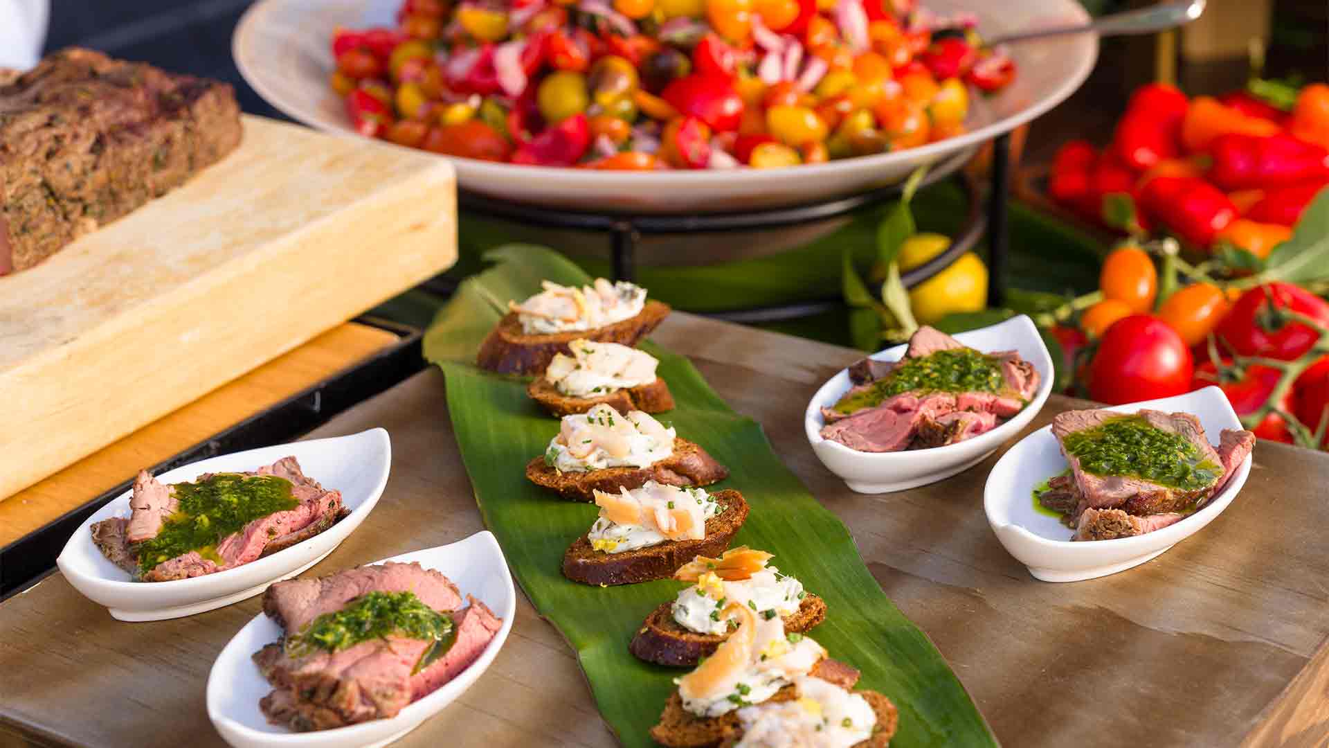 A rooftop catering spread featuring sliced steak with herb sauce, crostini bites, and fresh vegetables.