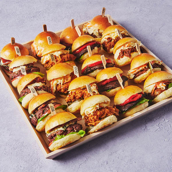 Assorted gourmet slider sandwiches arranged on a wooden catering tray, featuring mini beef burgers, crispy chicken sliders, and pulled chicken sandwiches on soft brioche buns.