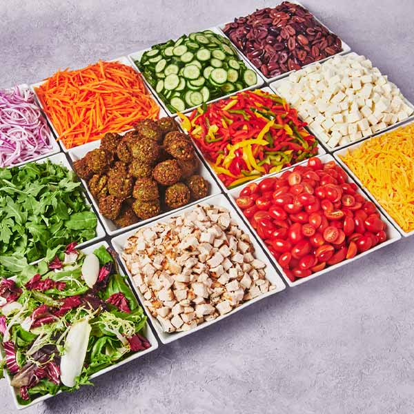 Catering salad bar spread with mixed greens, grilled chicken, falafel, cucumbers, carrots, peppers, tomatoes, feta cheese, beans, red onions, and shredded cheese.