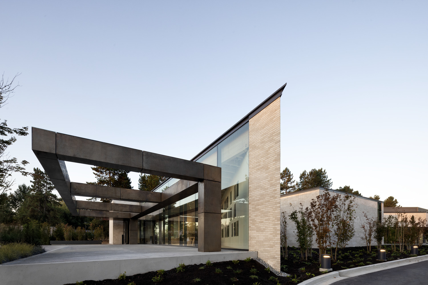 Modern building with large glass walls, light brick exterior, and a bold steel canopy, surrounded by landscaped greenery.